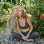 Full length positive sportswoman in gray leggings and sports bra sitting in lotus pose and kissing cute little son cheek after practicing yoga in green kush garden