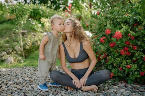Full length positive sportswoman in gray leggings and sports bra sitting in lotus pose and kissing cute little son cheek after practicing yoga in green kush garden