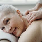 A senior woman with white hair smiles while receiving a therapeutic back massage indoors.