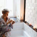 Woman in bathrobe enjoying aromatic candles for a serene bath experience.